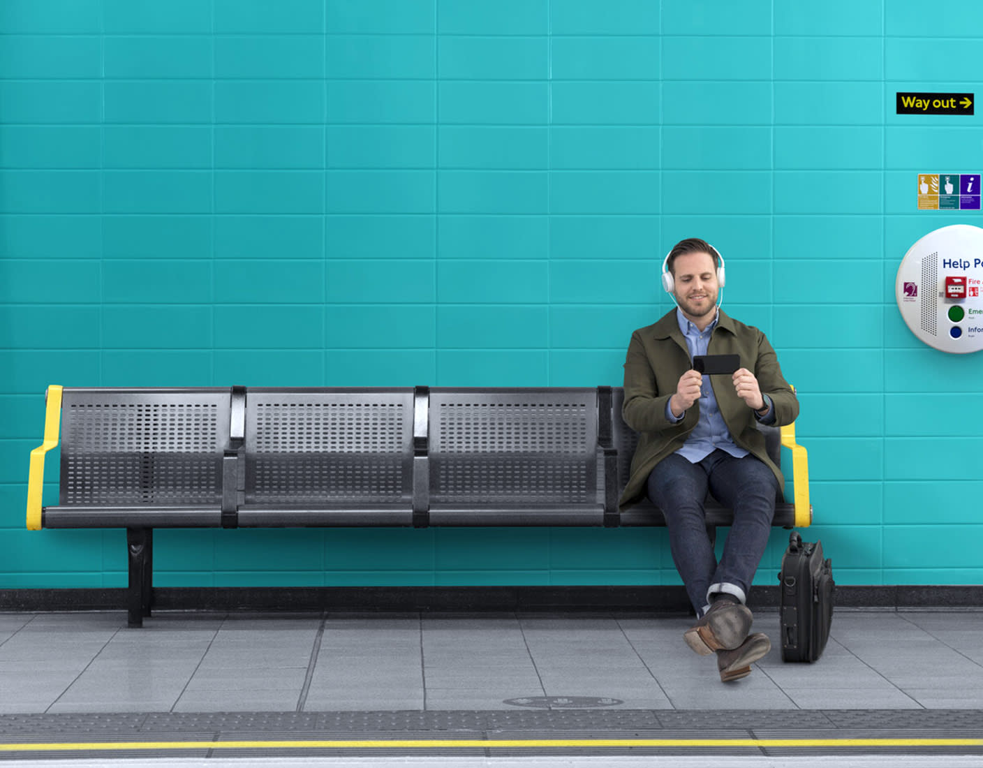 A man wearing headphones sits on a bench in a subway station, focused on his phone.